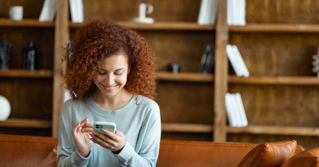 Smiling woman using a smartphone while sitting on a leather couch in front of a wooden bookshelf.